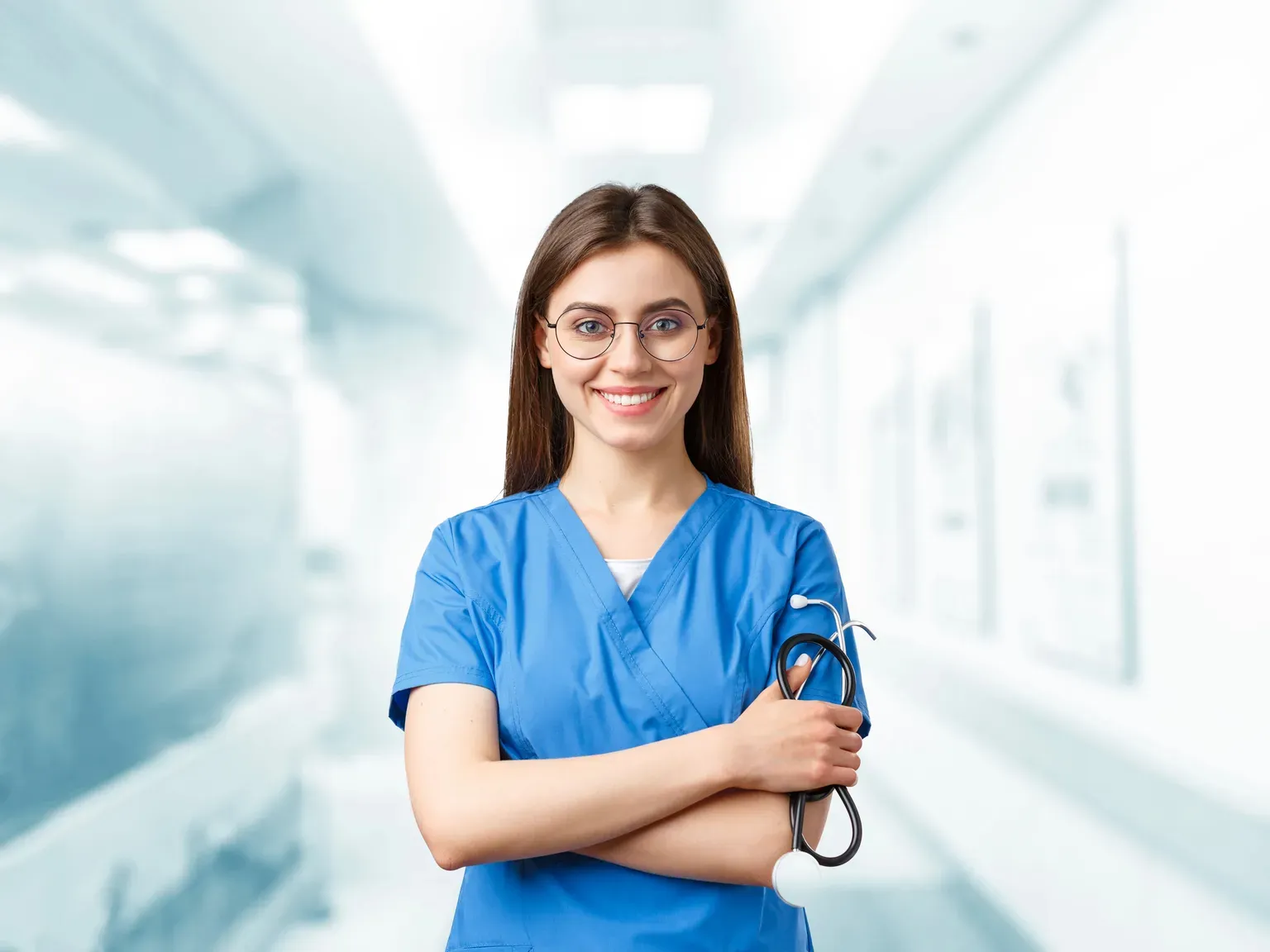 A smiling nurse practitioner student in blue scrubs holding a stethoscope in a bright hospital corridor.