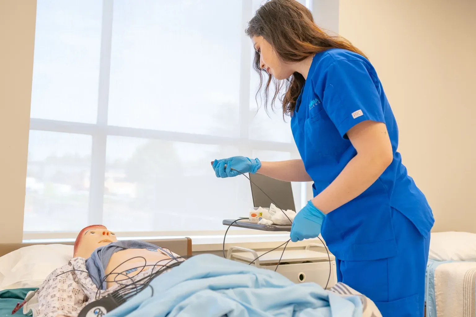A nurse in blue scrubs and gloves practicing a clinical procedure on a medical training mannequin in a simulation lab.